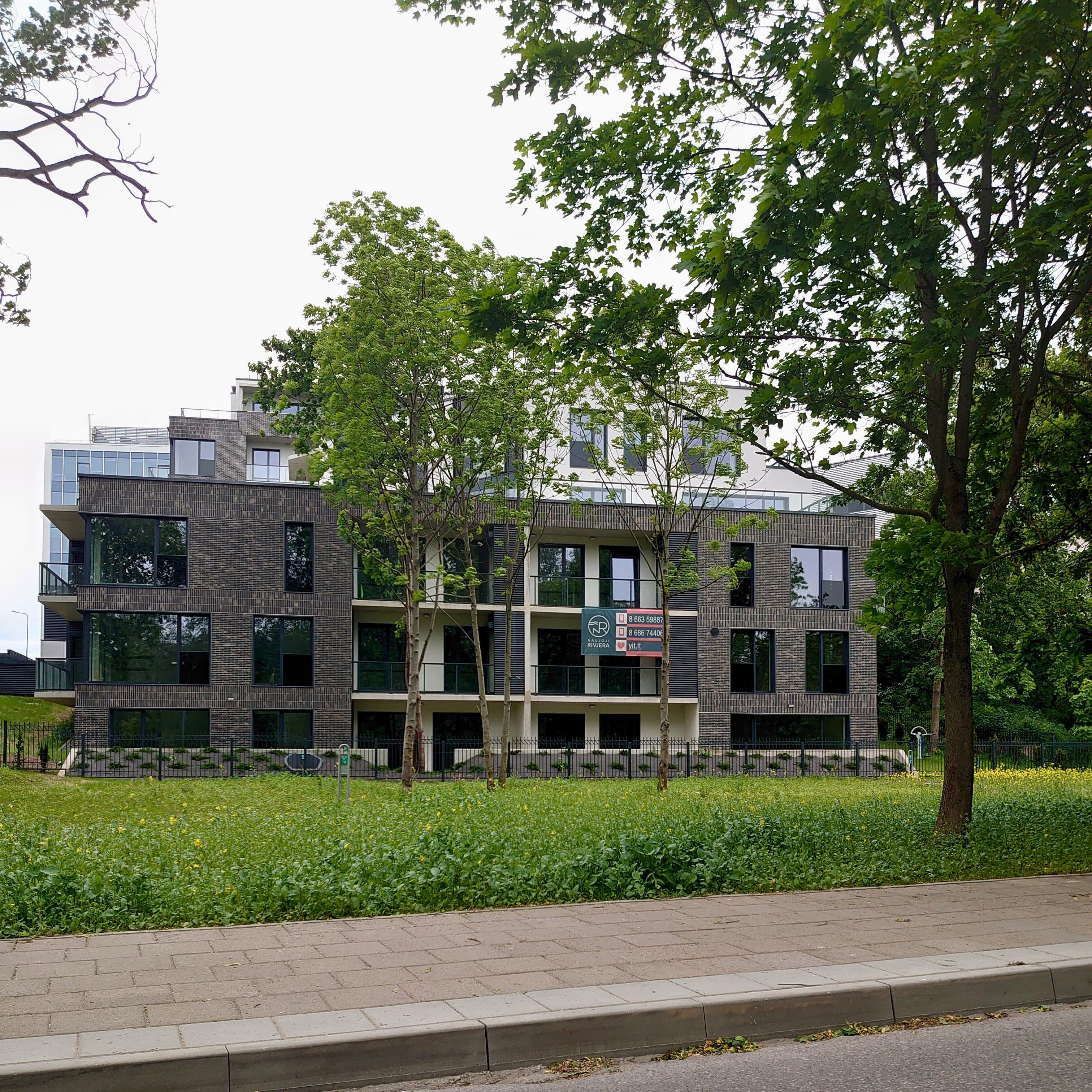 Didelis pastatas Lietuvoje su naujai sumontuotais langais, priekyje matyti medžiai.